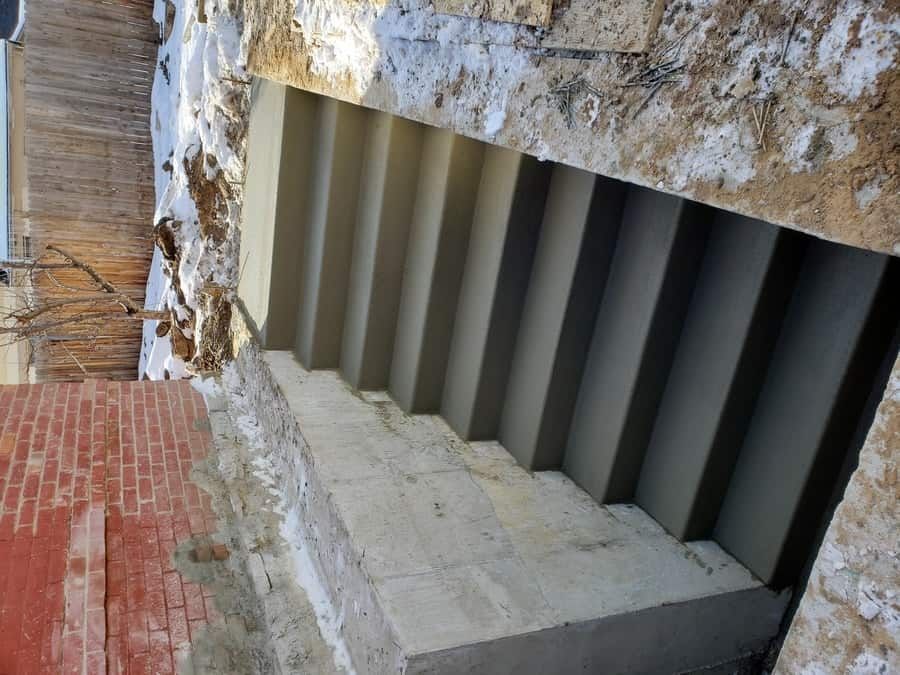 A set of newly poured gray concrete stairs leading down into a basement, adjacent to a brick wall and snow-covered ground.