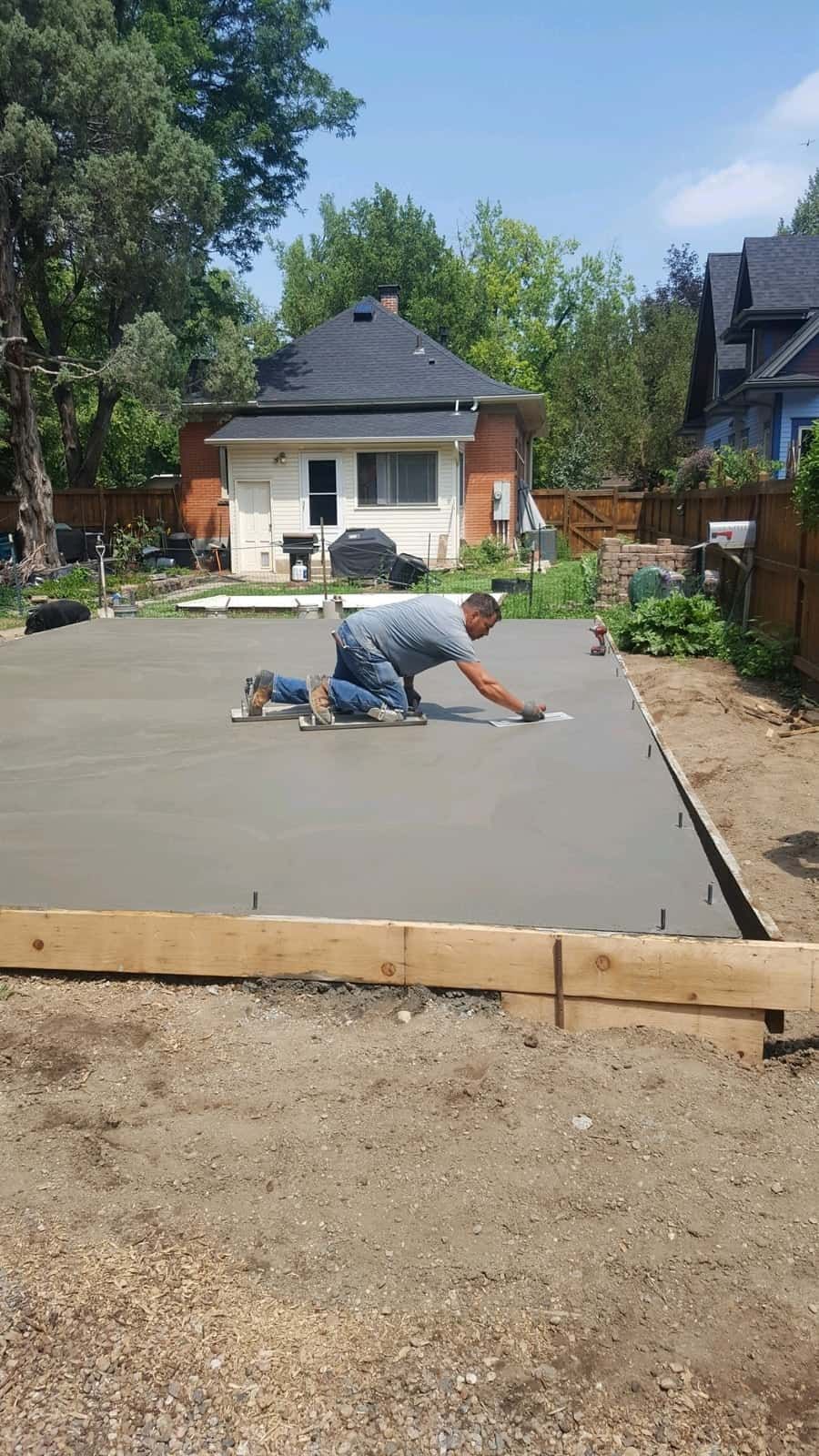A worker kneeling on a freshly poured concrete slab in a residential backyard, smoothing the surface with a tool.