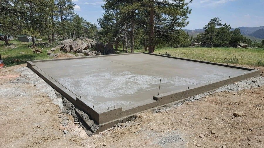 A fresh concrete slab foundation with anchor bolts installed, set in a rural, grassy landscape with pine trees.