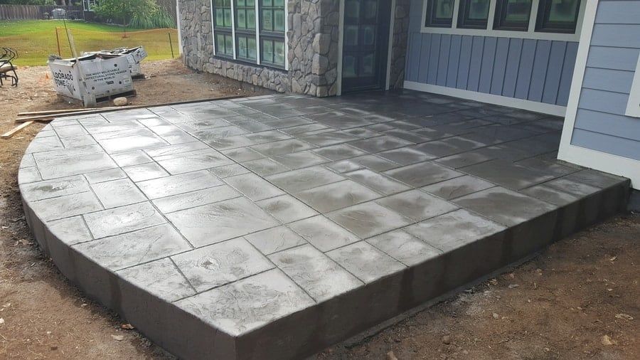 A newly installed grey concrete patio with a stamped tile pattern extends from the side of a house.