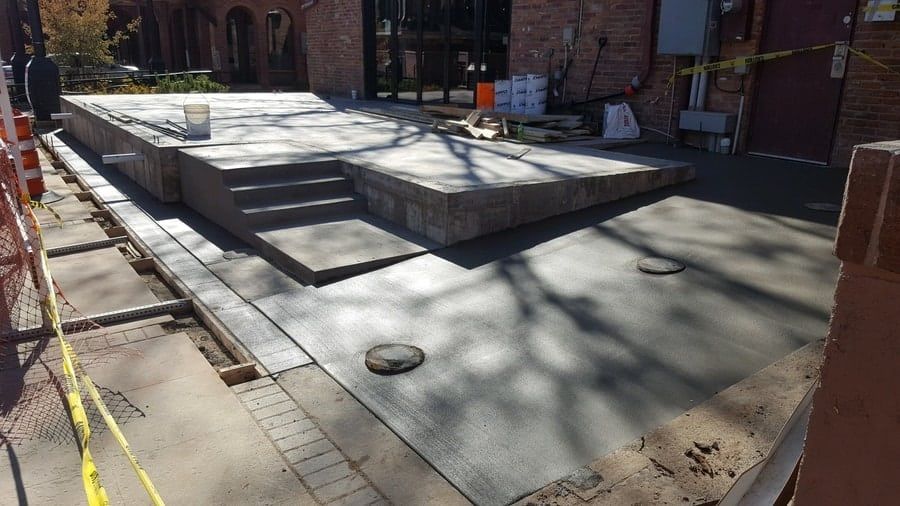 A newly poured concrete outdoor patio with a raised platform, steps, and a wheelchair ramp under construction.