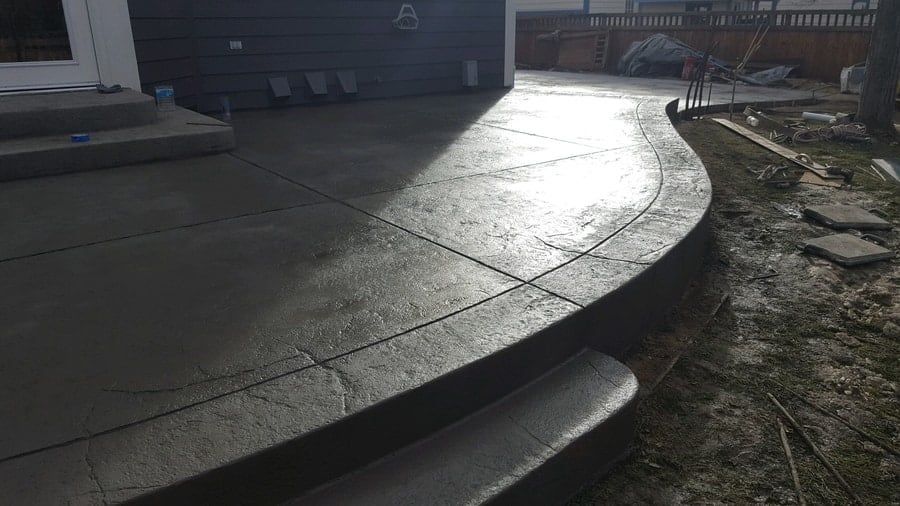 A newly poured concrete patio with a curved edge and a textured border extending from a residential home.