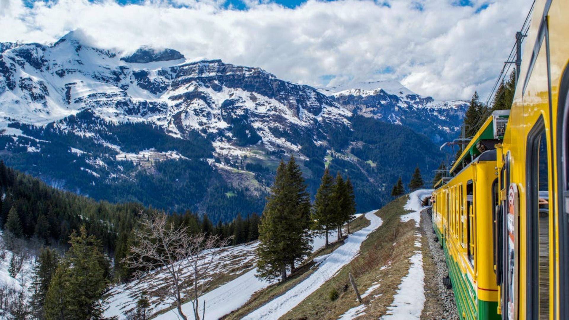 El tren amarillo viaja a lo largo de un sendero de montaña nevado; picos nevados al fondo.