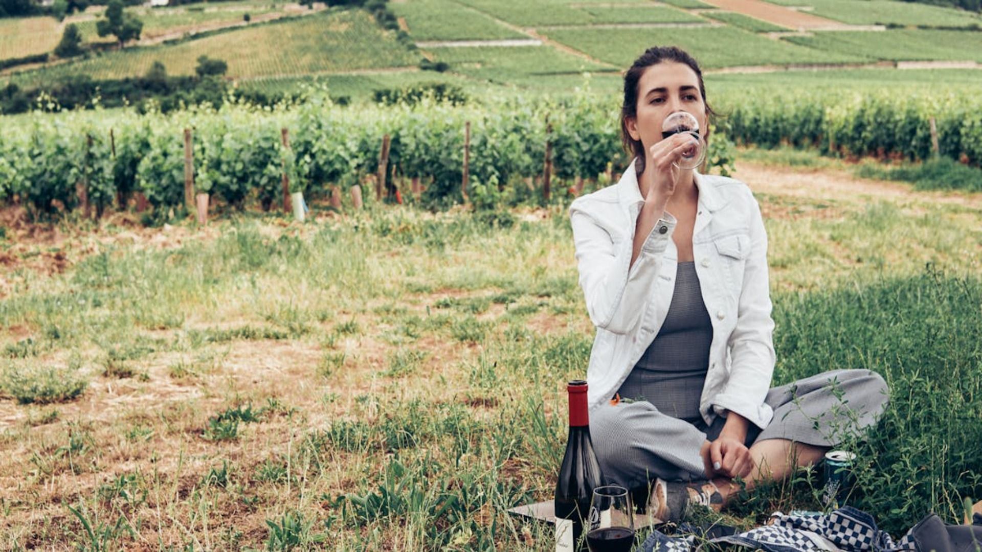 Una mujer sentada en un viñedo, bebiendo vino. Lleva una chaqueta blanca y un traje gris, y disfruta de la vista.