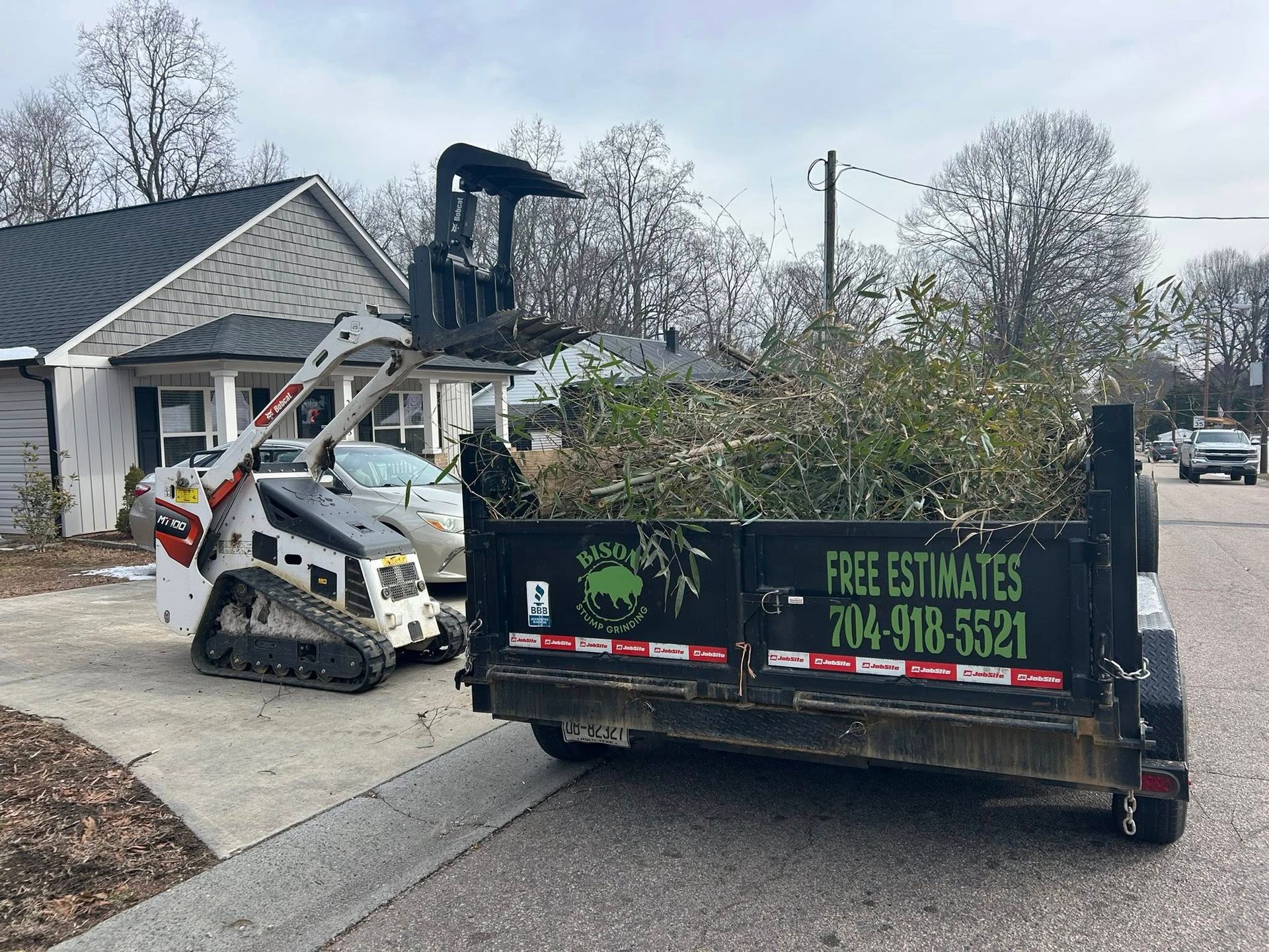Bobcat lifting debris into a dump trailer in a residential driveway beside a house.