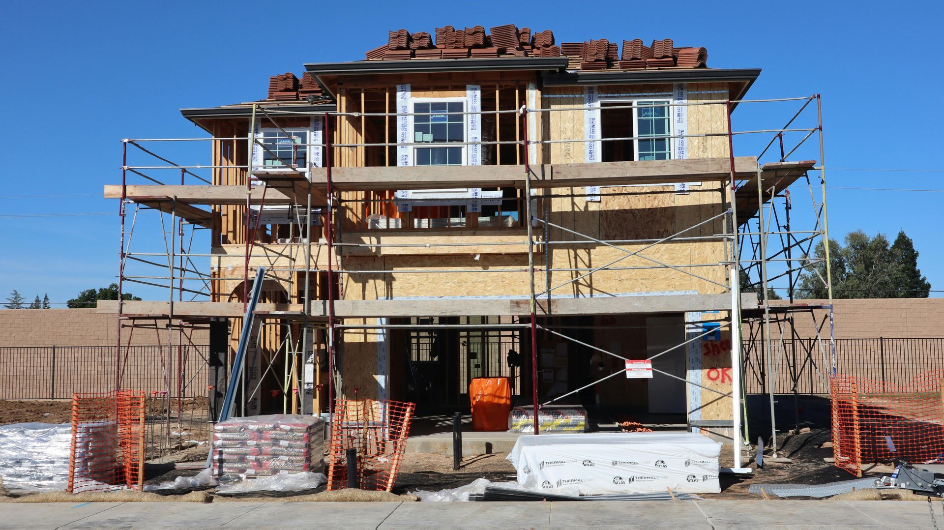 Three-story house under construction with scaffolding and rooftop work under a clear blue sky