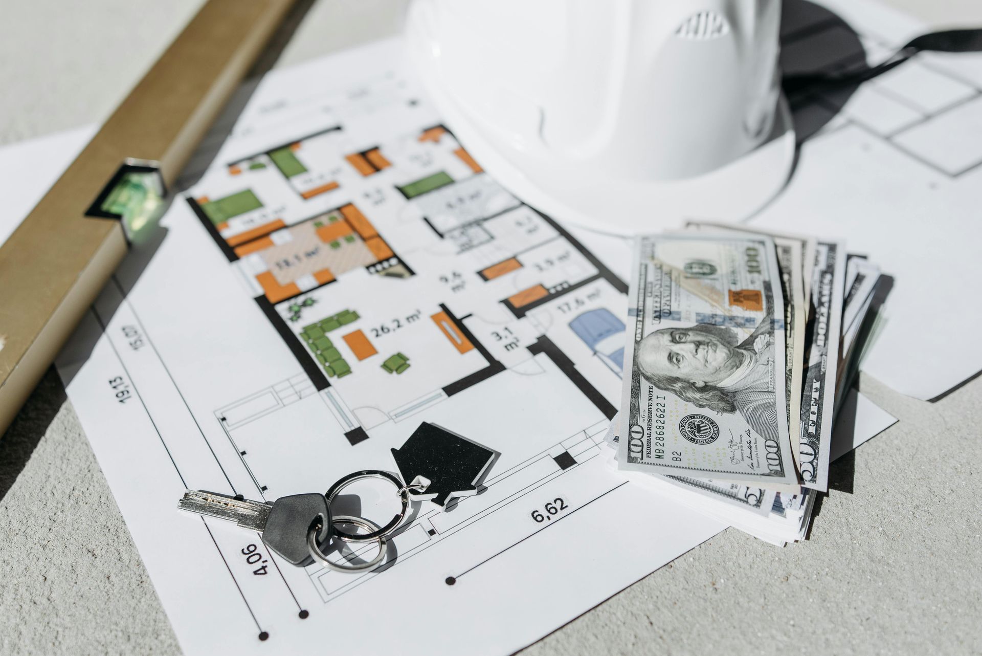 House floor plan with keys, rolled blueprint, and a stack of U.S. dollar bills on a desk