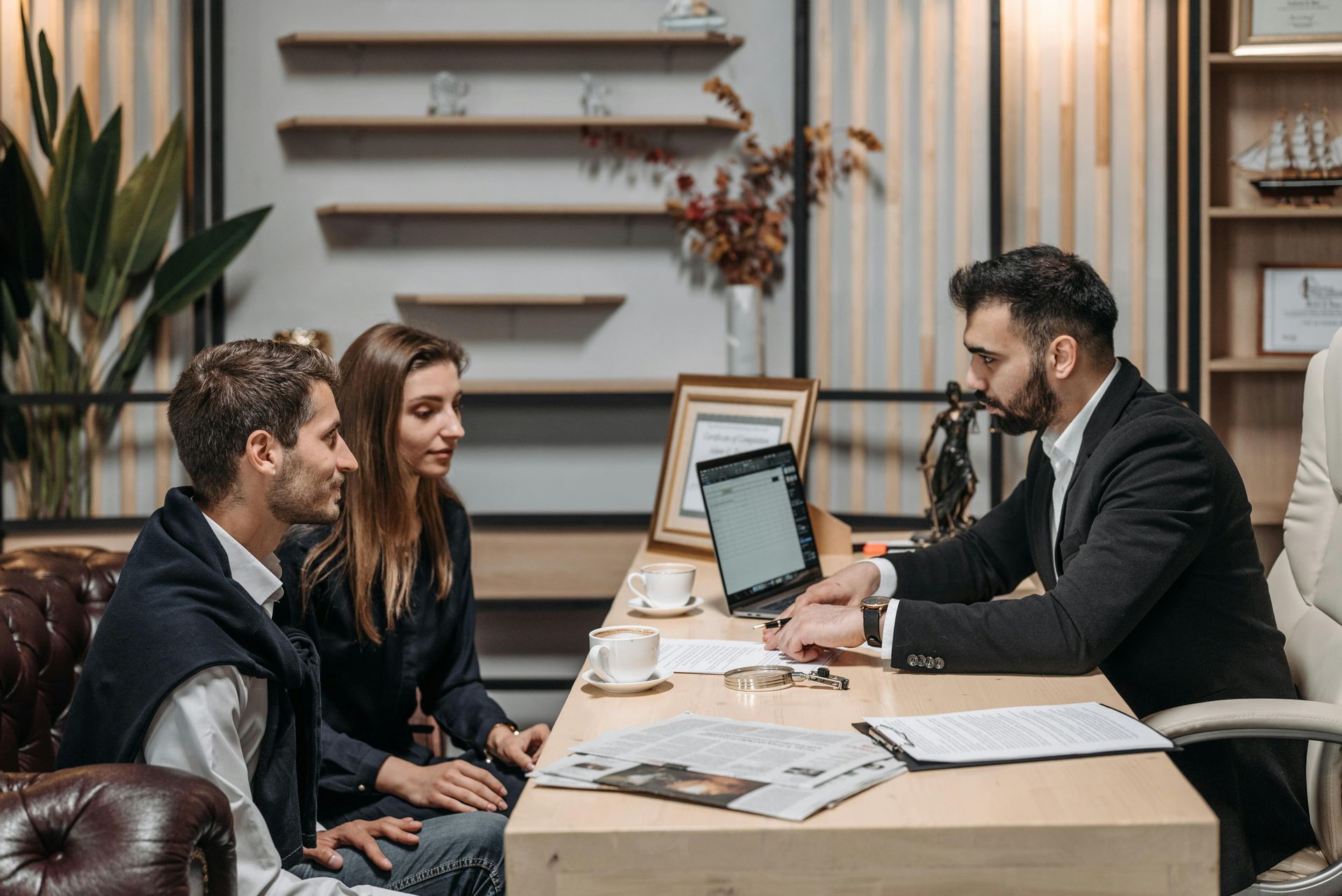 Three people in an office meeting across a desk, reviewing documents and a laptop, one man gesturing.