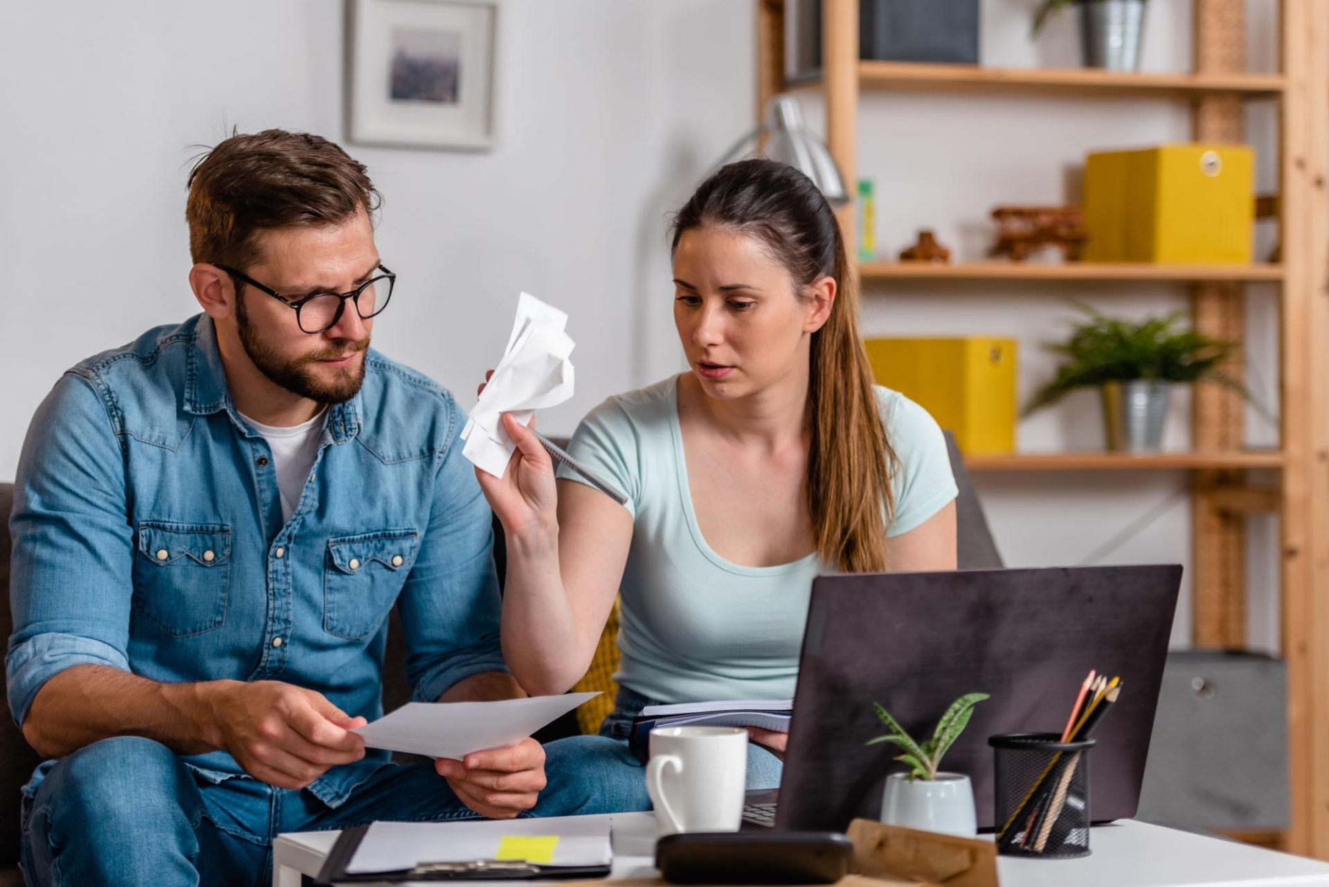 Two people reviewing bills at a table with a laptop, papers, and coffee mugs in a home office