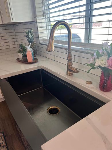 Stainless steel kitchen sink with gold faucet, white countertops, and a window.