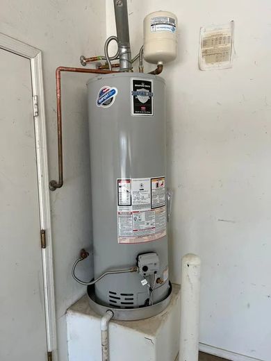 Water heater in a corner of a room, with copper pipes and white expansion tank.