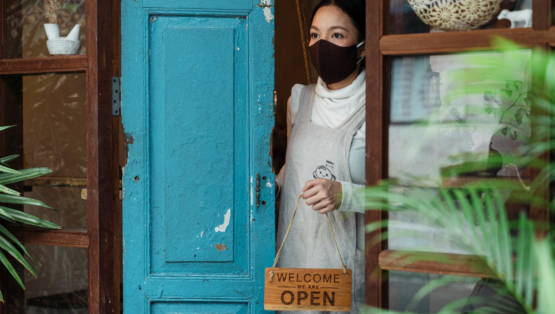 A woman wearing a mask is standing in front of a blue door holding a welcome sign.