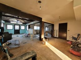 A living area undergoing renovation with exposed structural beams, light-colored walls, unfinished flooring, and tools.