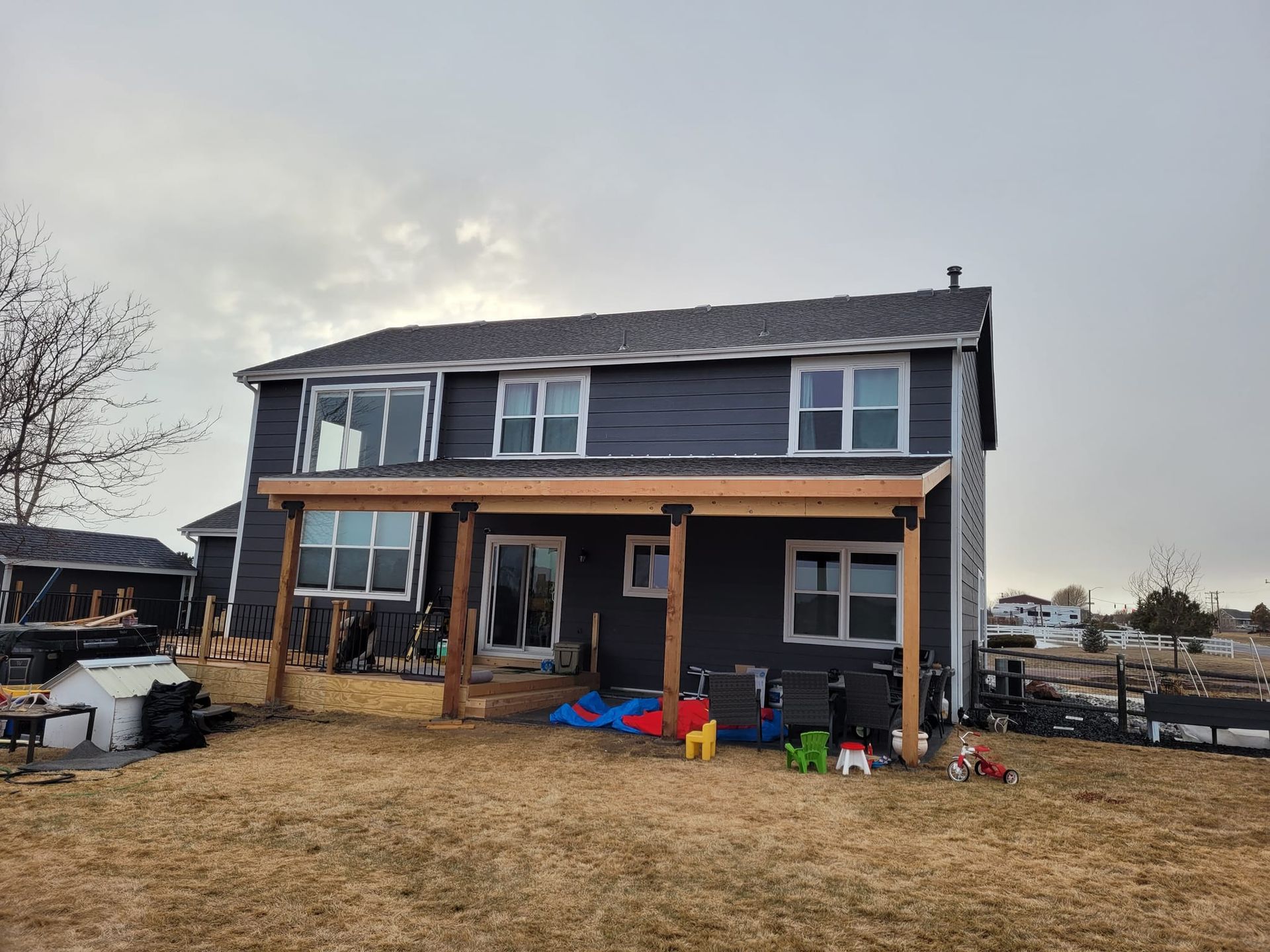 A two-story dark gray house with a newly constructed wooden porch roof over a patio on a dirt yard under a cloudy sky.