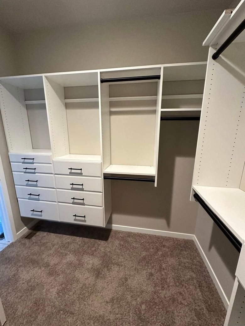 White custom closet storage system with built-in drawers and hanging rods against a light wall in a carpeted room.