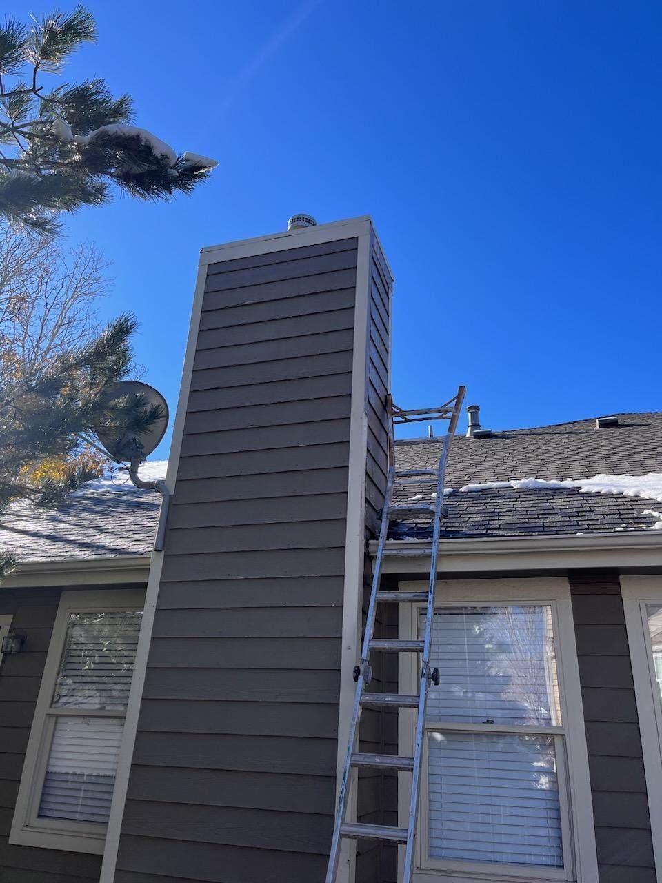 A ladder leans against a brown chimney on a house exterior with a snowy roof under a bright blue sky.