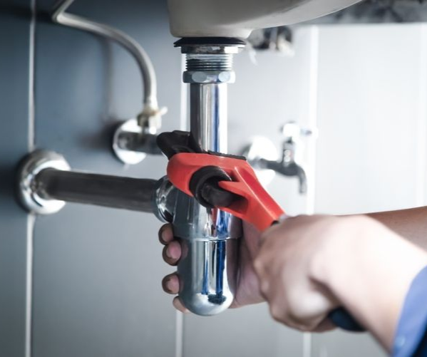 A plumber uses a red pipe wrench to tighten a chrome p-trap under a sink.