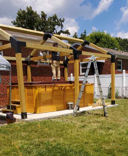 Construction of a wooden pergola over a hot tub in a backyard. A person stands on the hot tub frame.