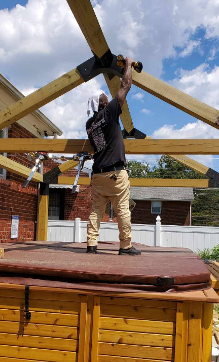 Man on hot tub roof, using power tool to assemble wooden patio structure. Sunny day.