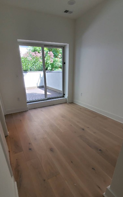 Empty room with hardwood floors, sliding glass door to balcony. White walls, natural light.