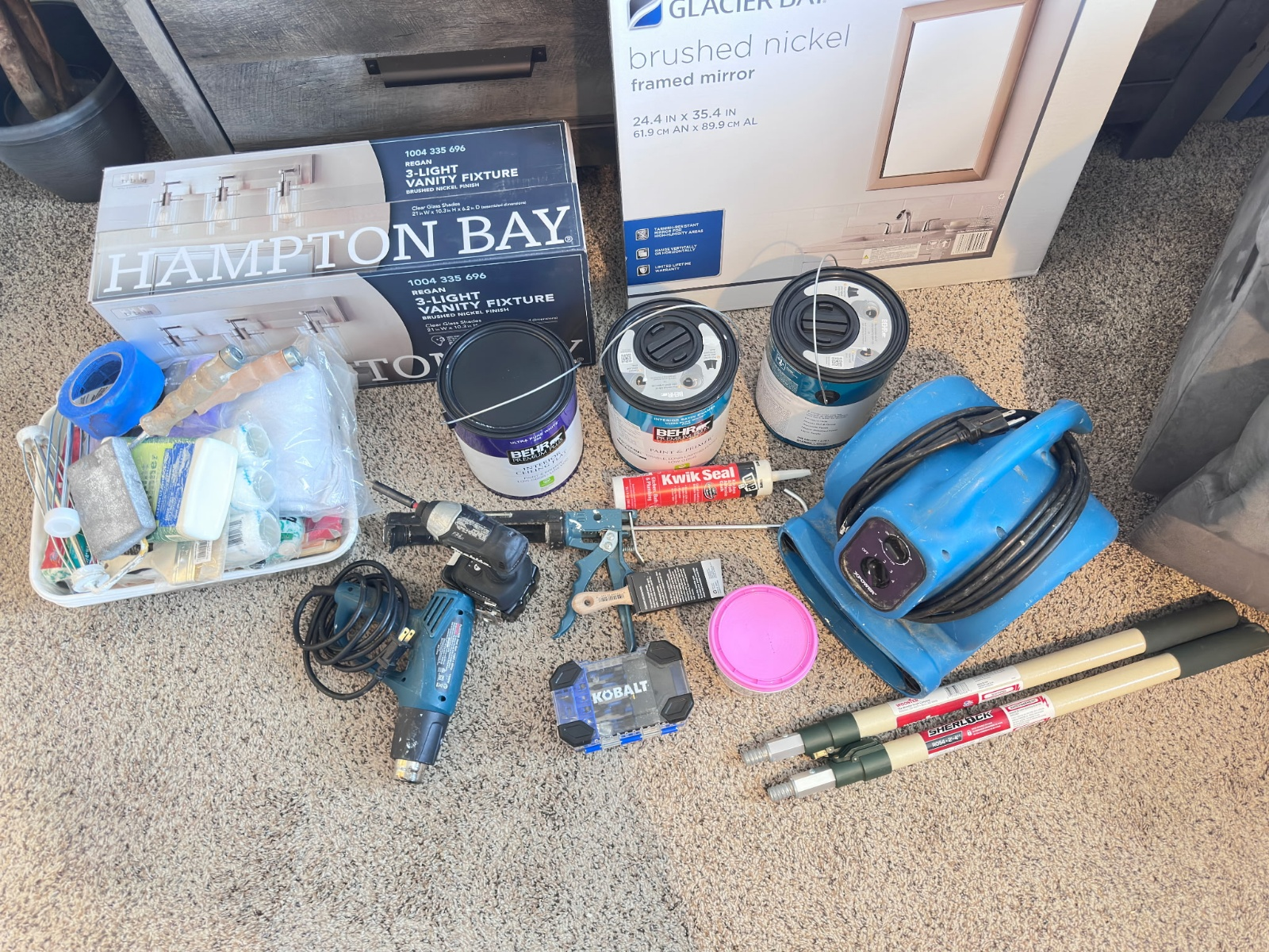 Assorted painting supplies on a carpeted floor, including paint cans, brushes, tools, and a Hampton Bay box.
