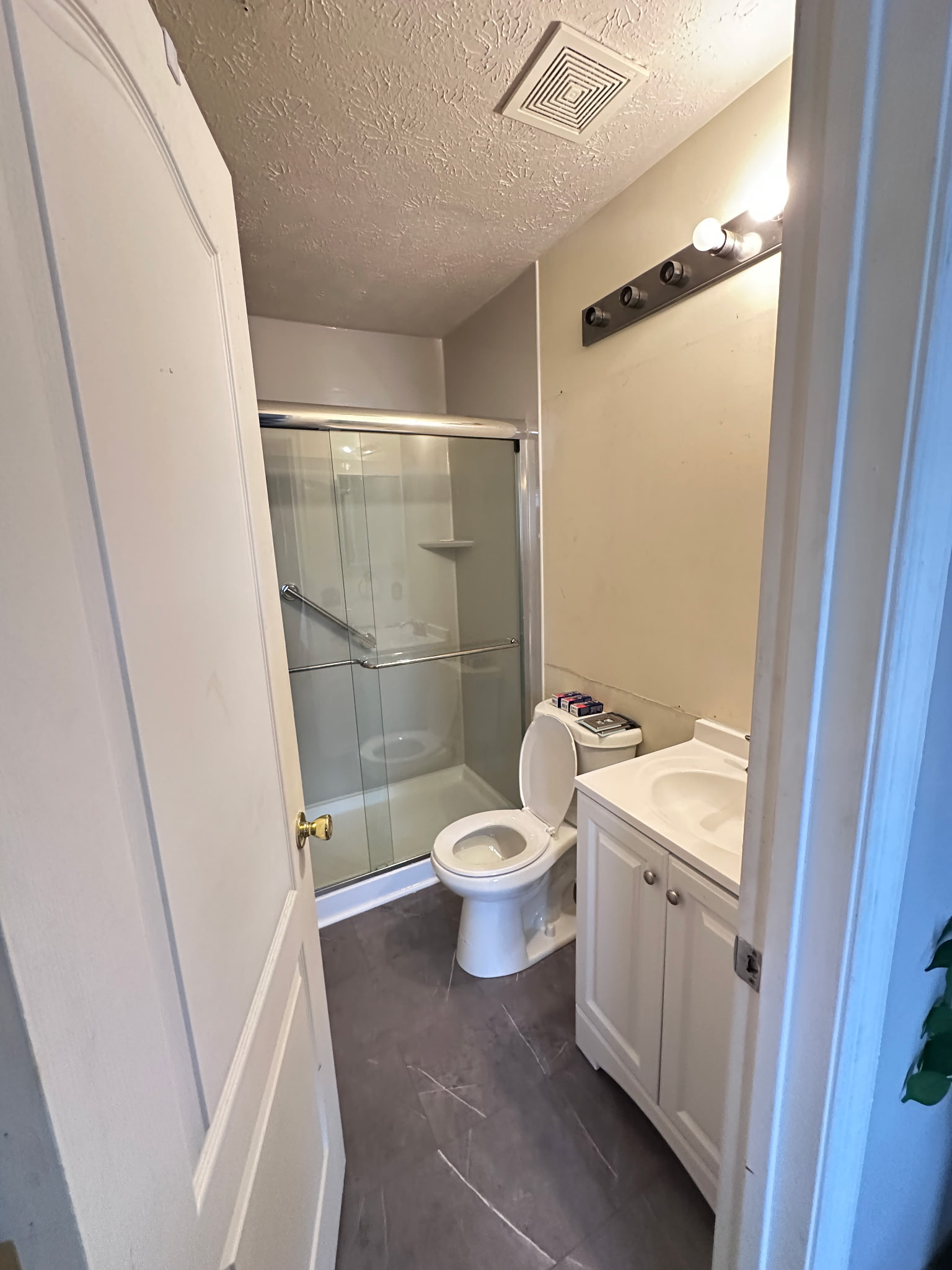 Small bathroom with a toilet, shower, and vanity. Gray flooring and light walls.