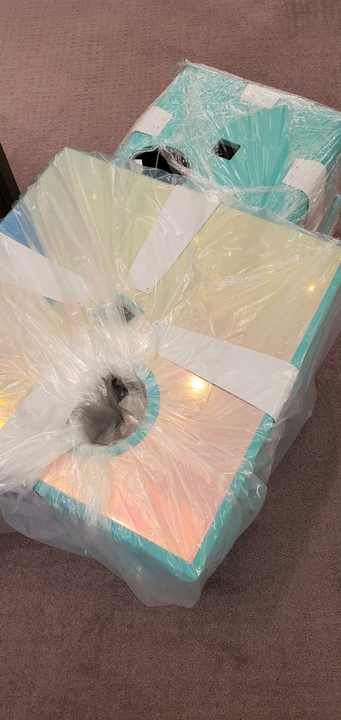 Boxes wrapped in plastic, stacked, with iridescent surfaces in shades of pink, yellow, and blue.