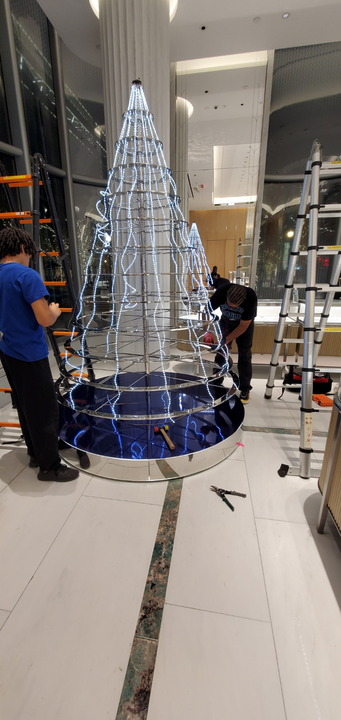 Workers installing a lighted cone-shaped structure in a building. The structure has a blue base and silver frame.