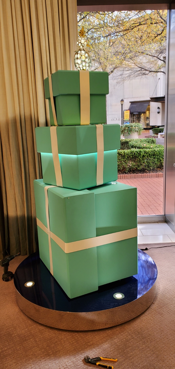 Stacked teal gift boxes with white ribbons on a circular platform, near a window.