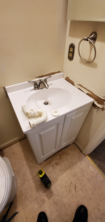 A small, white bathroom sink cabinet.  A partially installed sink with plumbing and a towel ring are visible. 