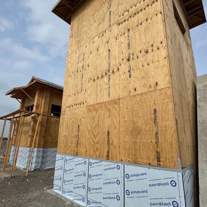 Two wood-framed structures under construction, sheathed in plywood and wrapped with protective material at the base.