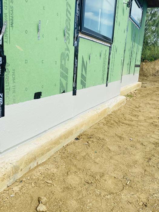 Exterior view of a building under construction, showing green sheathing, windows, and foundation against a dirt ground.