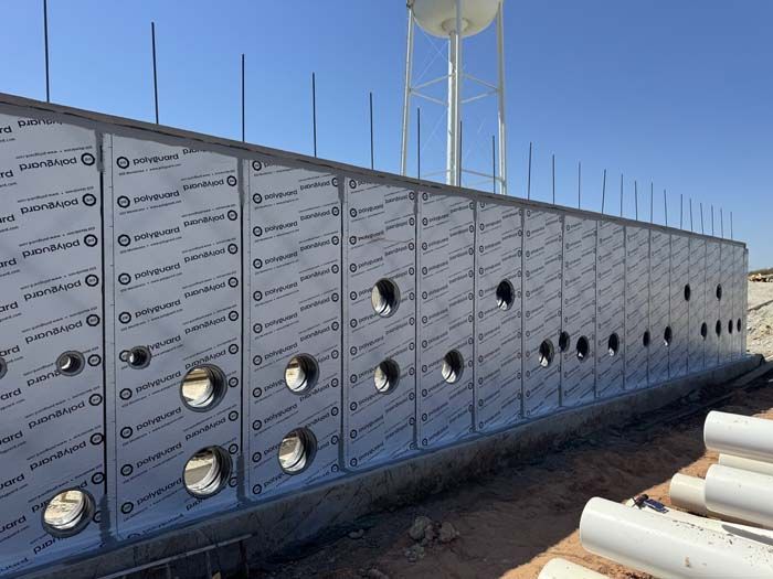 Exterior wall under construction, silver panels with circular openings. Water tower in background.