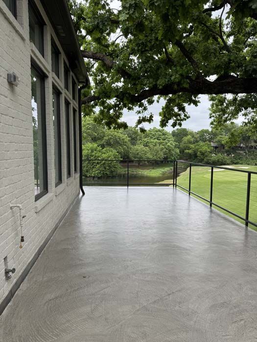 Gray concrete patio next to a white brick building, overlooking a grassy area and water.