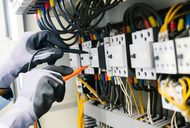 Hands in black gloves testing electrical wiring in a control panel.
