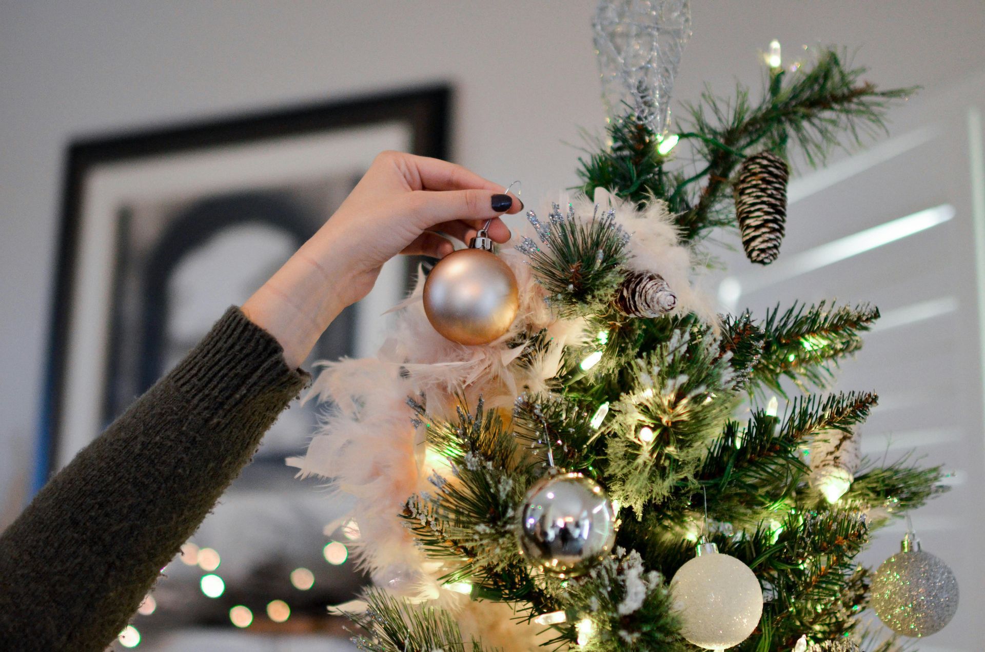Person hangs a gold ornament on a decorated Christmas tree with lights, feathers, and other ornaments.