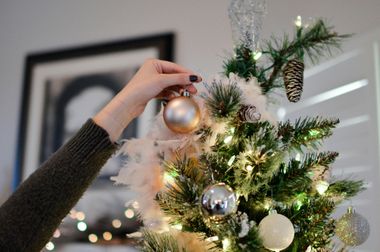 Person hangs a gold ornament on a decorated Christmas tree with lights, feathers, and other ornaments.
