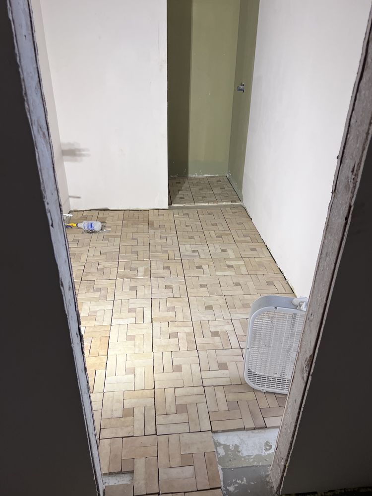Room with beige rectangular floor tiles, partially installed. White walls, bucket visible.