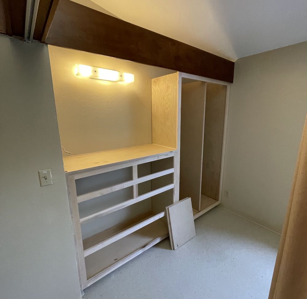 Built-in closet with shelves and drawers under a light fixture in a room with beige walls and a low ceiling.