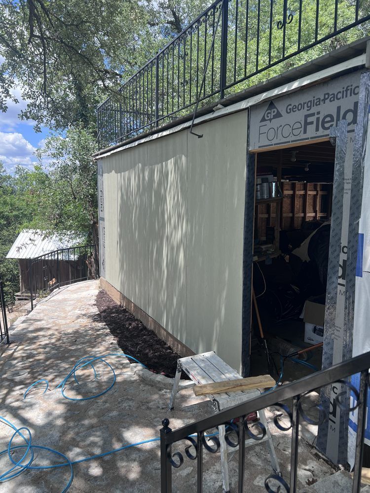 Exterior of a building under construction, with siding and an open doorway. Black metal railing and a pathway.