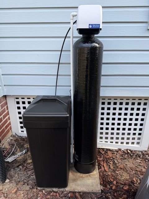 Water softener system outside a house, with a black tank, a salt tank, and white control unit.
