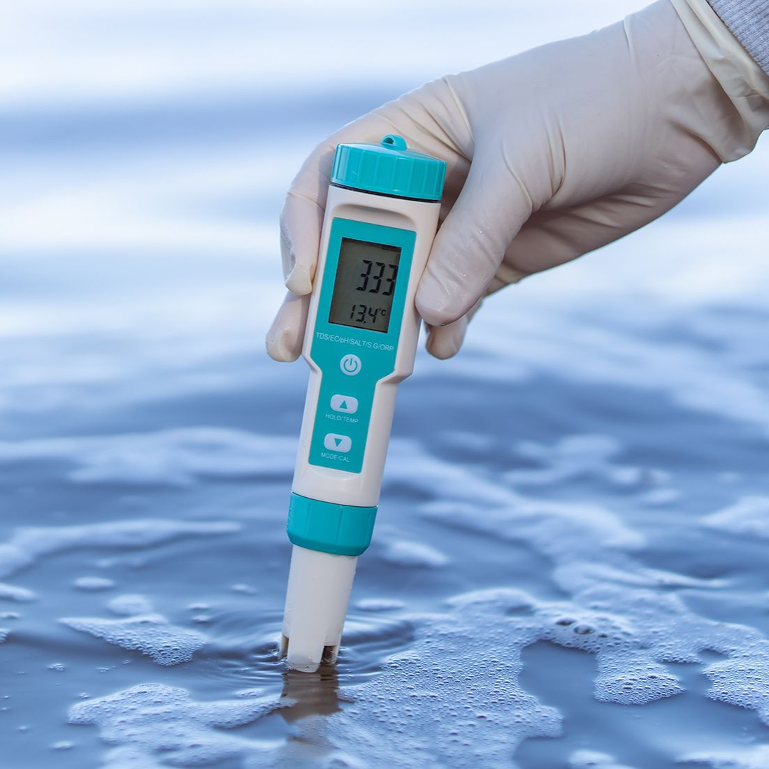 Gloved hand holding a digital meter in ocean water, measuring. Display shows readings on the screen.