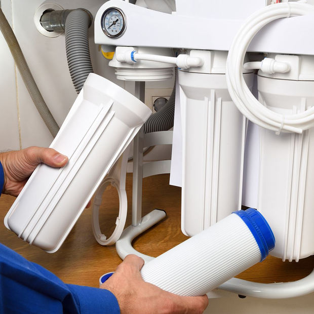 Person changing water filter cartridges under a sink; white cartridges and filter system.