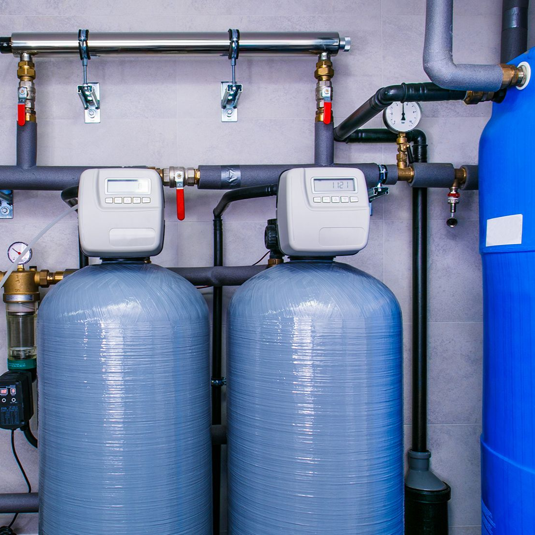 Water filtration system with two gray tanks, gauges, and pipes in a utility room.