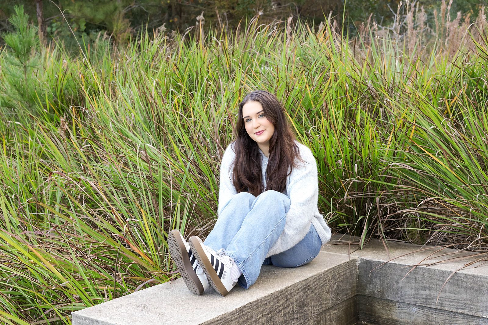 A smiling person sits on a stone ledge in front of tall green grasses, wearing a white hoodie, blue jeans, and sneakers.