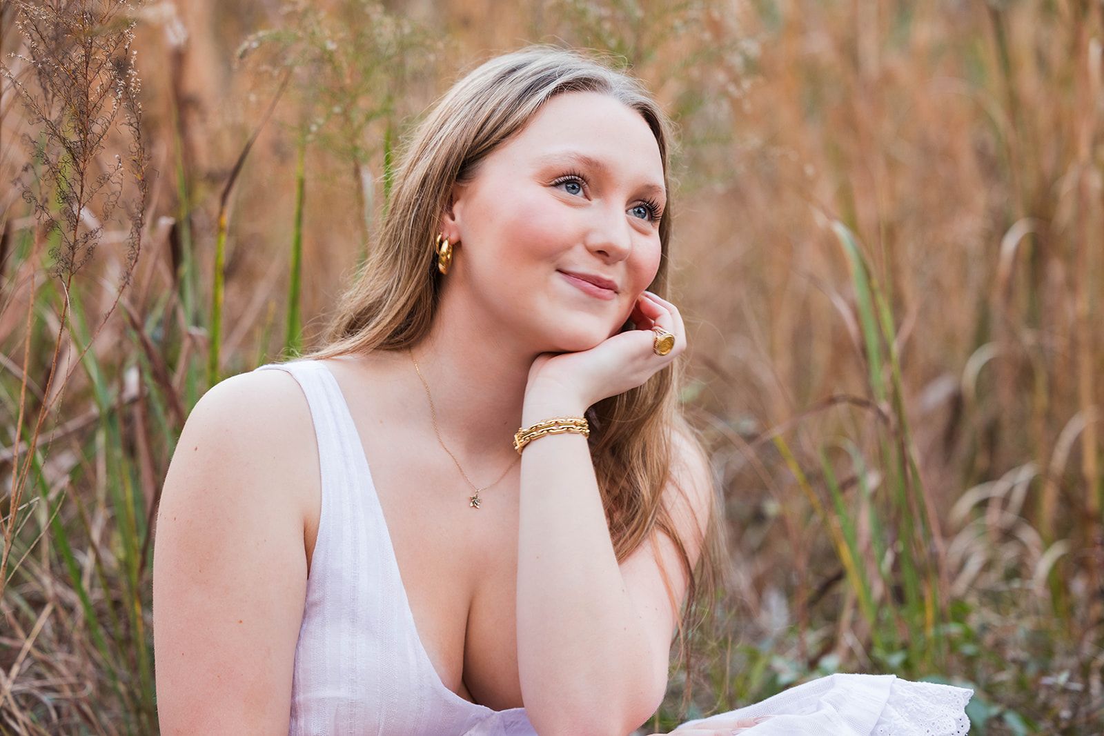 A person with long hair rests their chin on their hand, smiling softly while sitting in a field of tall, dry grass.