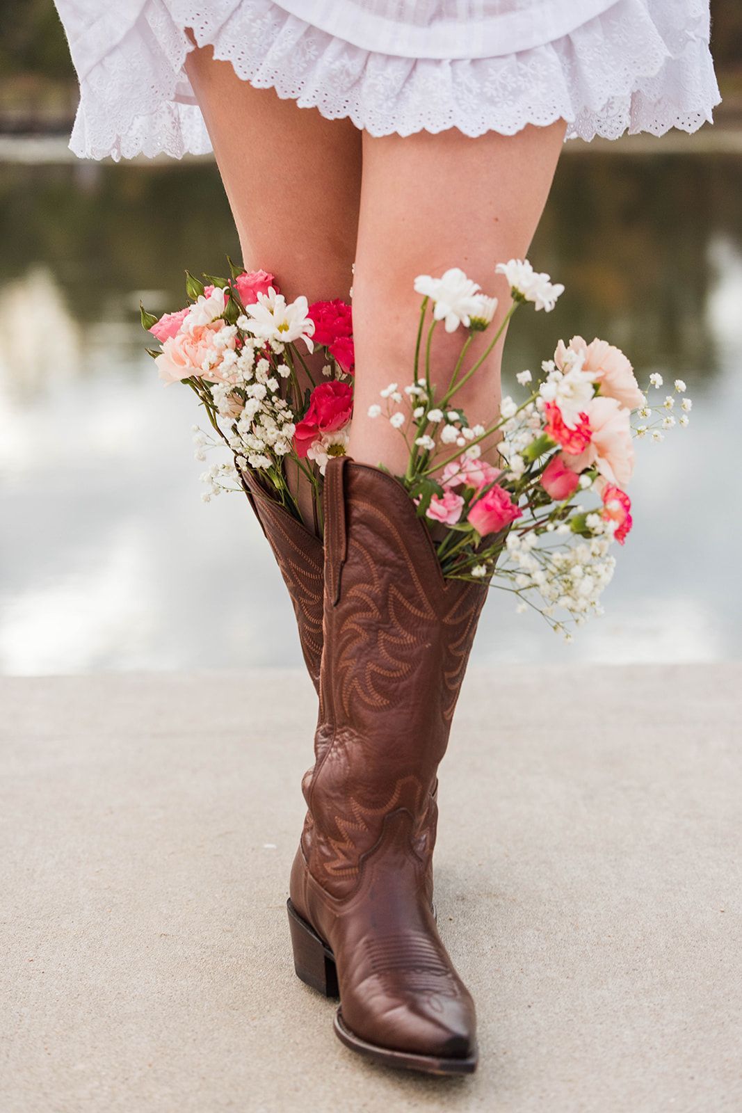 A pair of brown cowboy boots filled with white and pink flowers worn with a white dress outdoors.