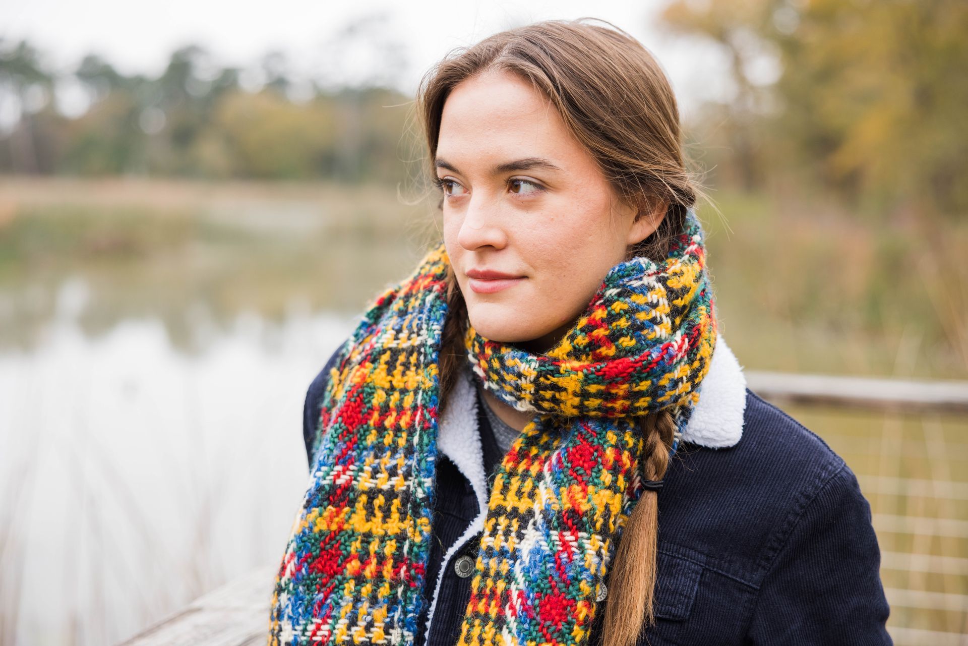 A person wearing a thick, multicolored plaid scarf and a dark coat stands outdoors near a calm body of water.