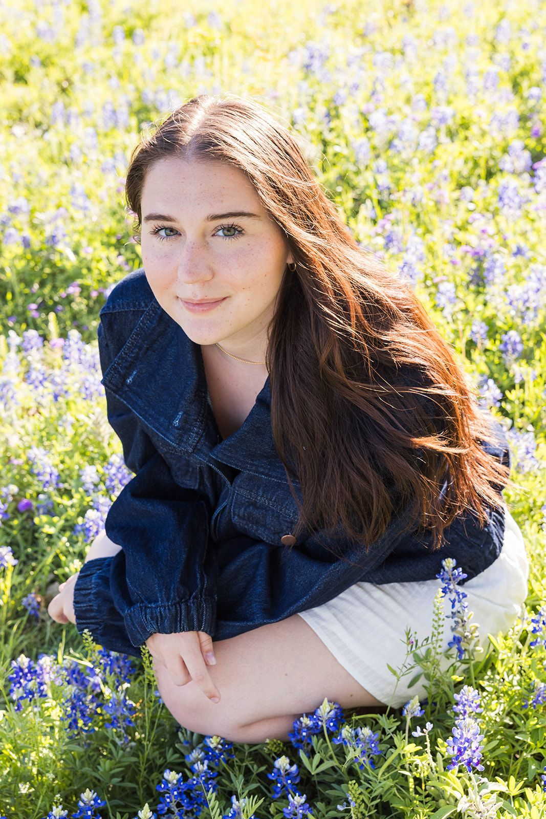 A person in a blue denim jacket squats in a vibrant field of bluebonnet flowers, smiling toward the camera.
