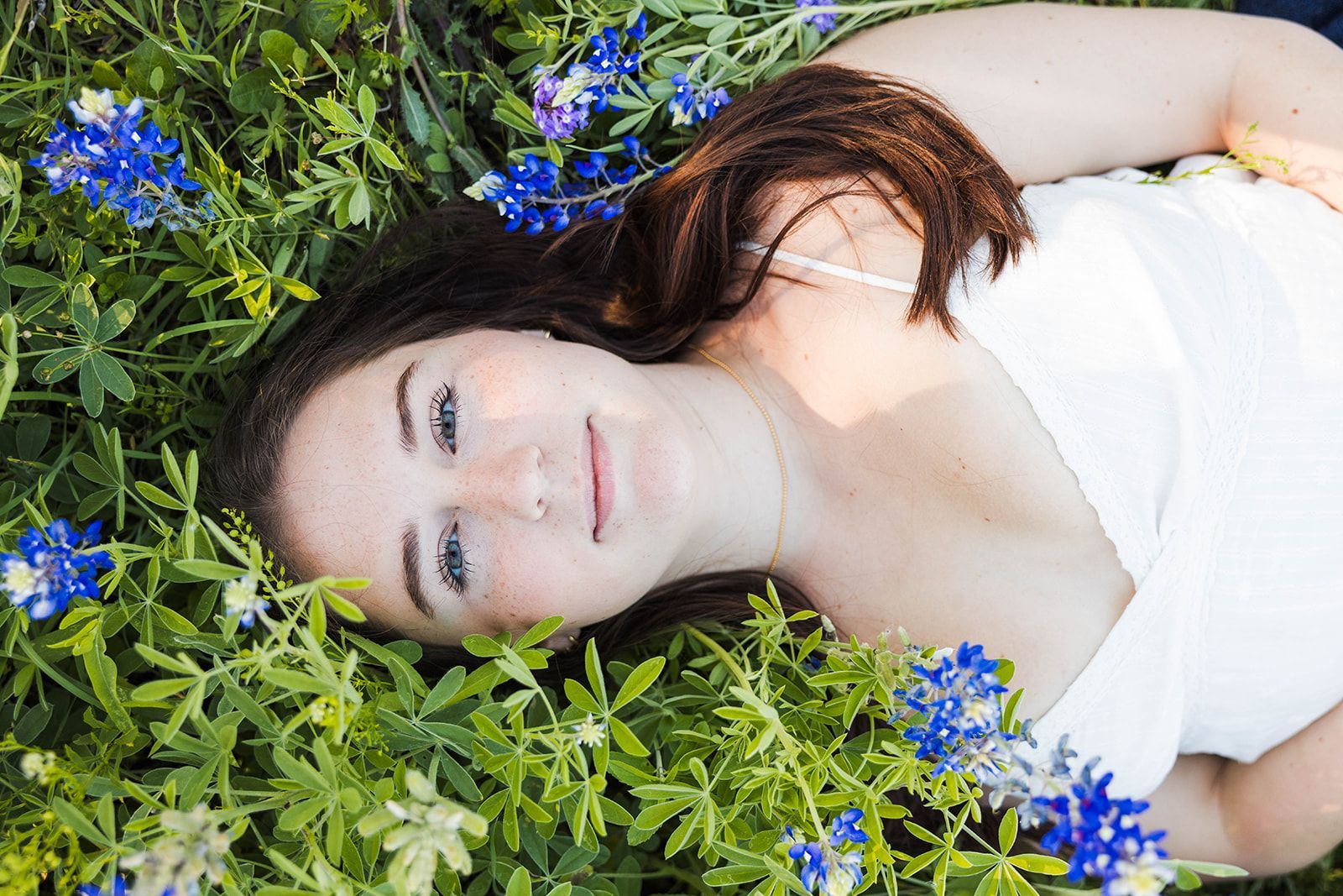 A person with dark hair wearing a white top lying in a field of bluebonnet flowers and looking upward.